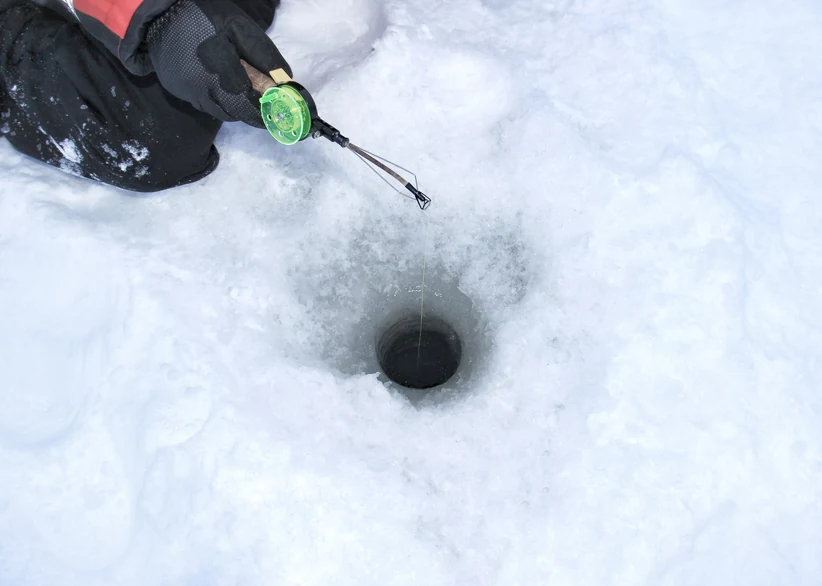 A gloved hand holds a short ice fishing rod with a bright green reel over a round hole drilled in the snow-covered ice, with the fishing line dropping into the dark water below.