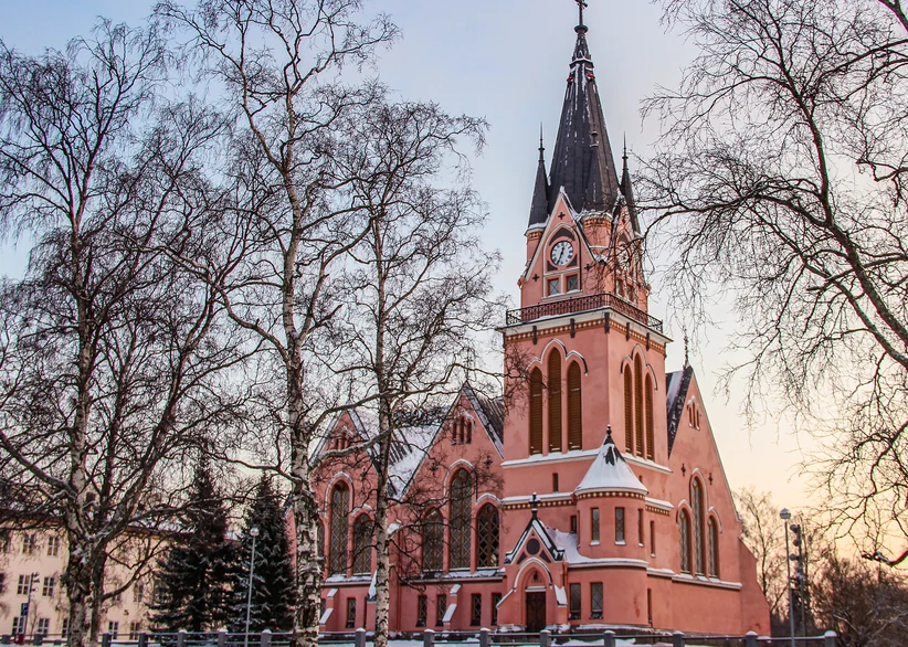 The red-brick Kemi Church with its tall dark spire and clock tower stands among bare birch trees in winter, lit by soft pink dusk light.