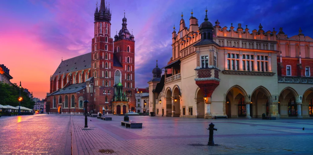 St. Mary's Basilica with its two asymmetrical towers and the Renaissance Cloth Hall glow at dawn on Kraków's Main Market Square, beneath a vivid pink and purple sky with wet cobblestones reflecting the light.