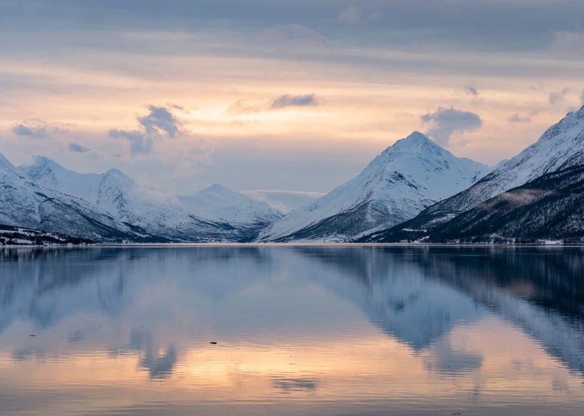 Snow-capped peaks rise sharply above the calm waters of Lyngenfjord, their reflection mirrored perfectly on the still surface beneath a soft pink and orange winter sunset sky.