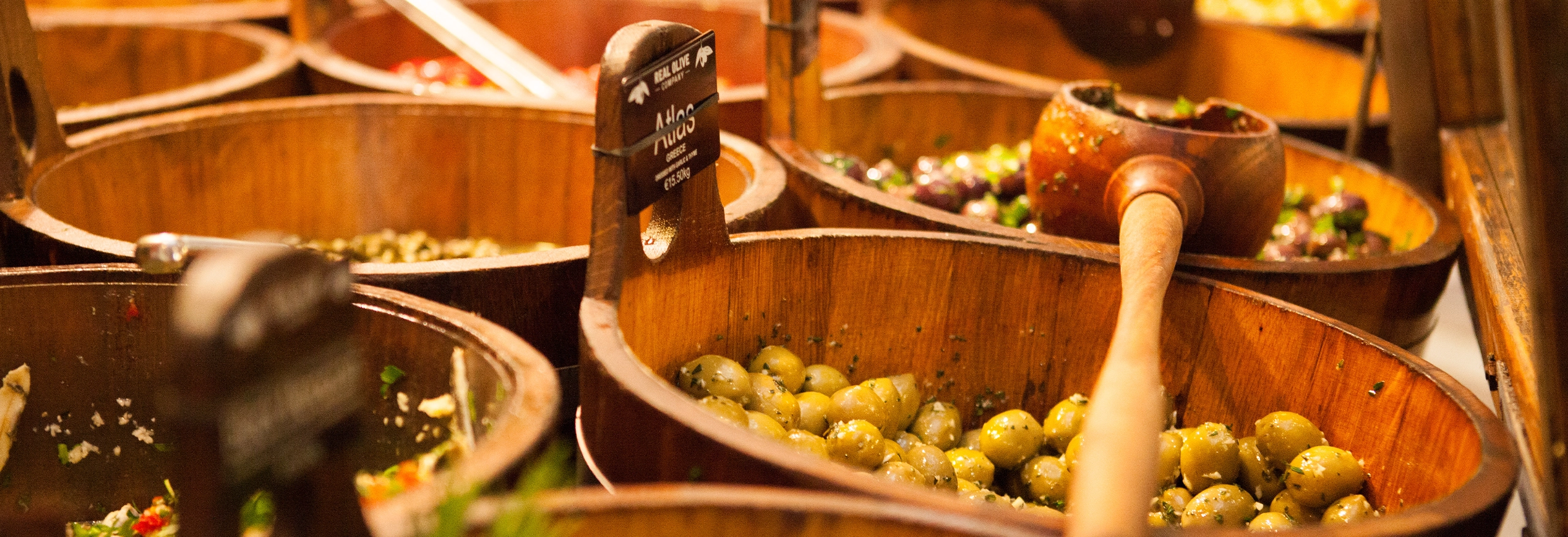 Wide view of wooden olive barrels and serving scoops on a market stall counter.