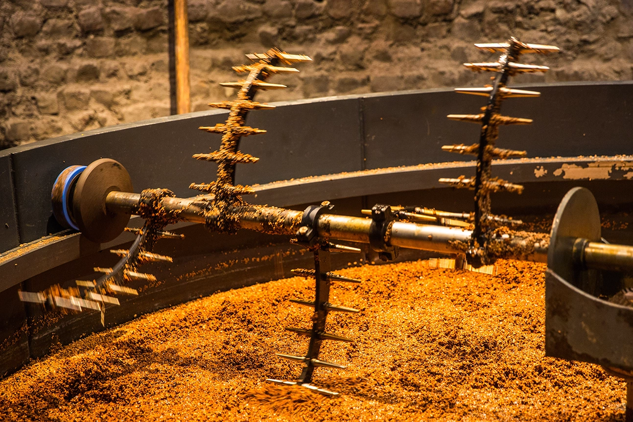 Metal rake arms sweep through coarse golden malted grain inside a large copper-lit mash tun.