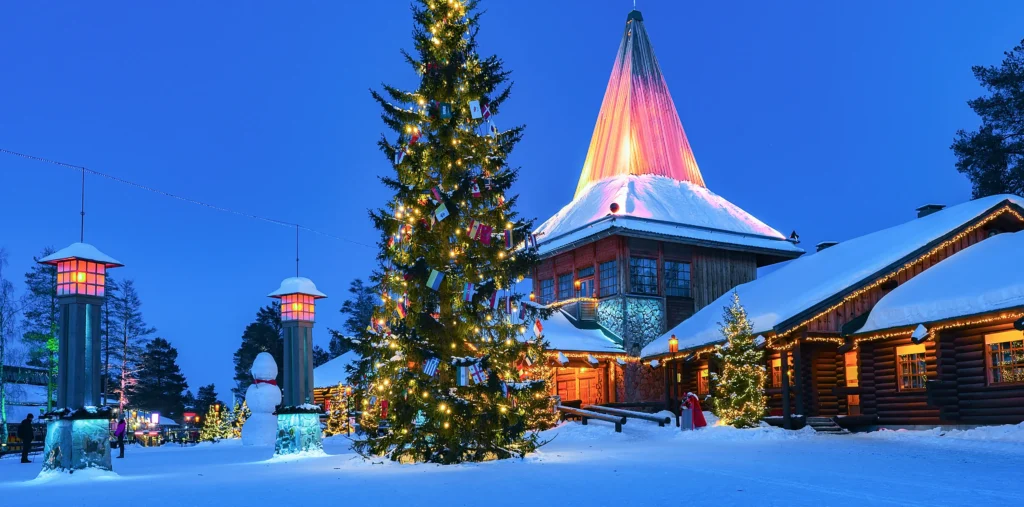 Santa Claus Village in Rovaniemi at blue-hour dusk, with a tall Christmas tree strung with lights and flags, glowing red lanterns, a snowman, and snow-covered log cabins beside an illuminated pointed tower.