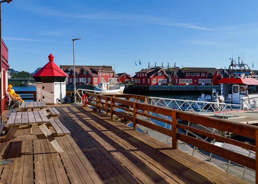 A sunlit wooden pier with picnic tables and a red-roofed lantern overlooks a harbor lined with traditional red fishing cabins and moored boats under a clear blue sky.