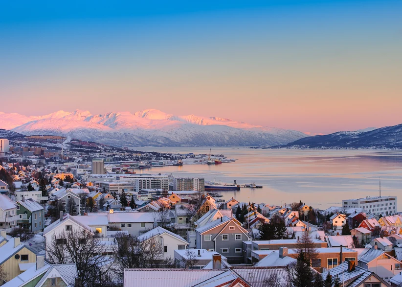 A panoramic view of snow-covered rooftops in Tromsø overlooking a calm fjord, with pink-tinted snow-capped mountains glowing under a soft sunset sky.