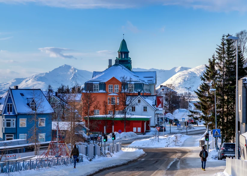 A snowy street in Tromsø lined with colorful wooden buildings leads toward the historic orange town hall with its green domed tower, framed by snow-capped mountains.