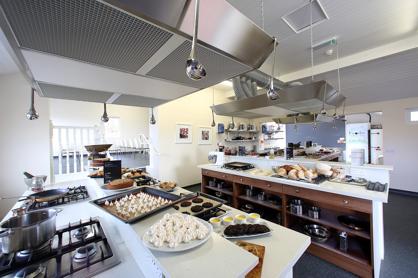 Bright demonstration kitchen with gas hobs, meringues, cakes and fresh breads laid out on white counters.